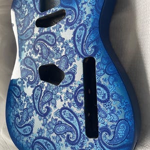 K&ouml;nnte beinhalten: Ein blauer Gitarrenkorpus mit einem Paisley-Muster in Blau- und Wei&szlig;t&ouml;nen. Der Korpus hat Aussparungen f&uuml;r Tonabnehmer und andere Hardware. Die Oberfl&auml;che ist gl&auml;nzend, und das Design ist detailliert.