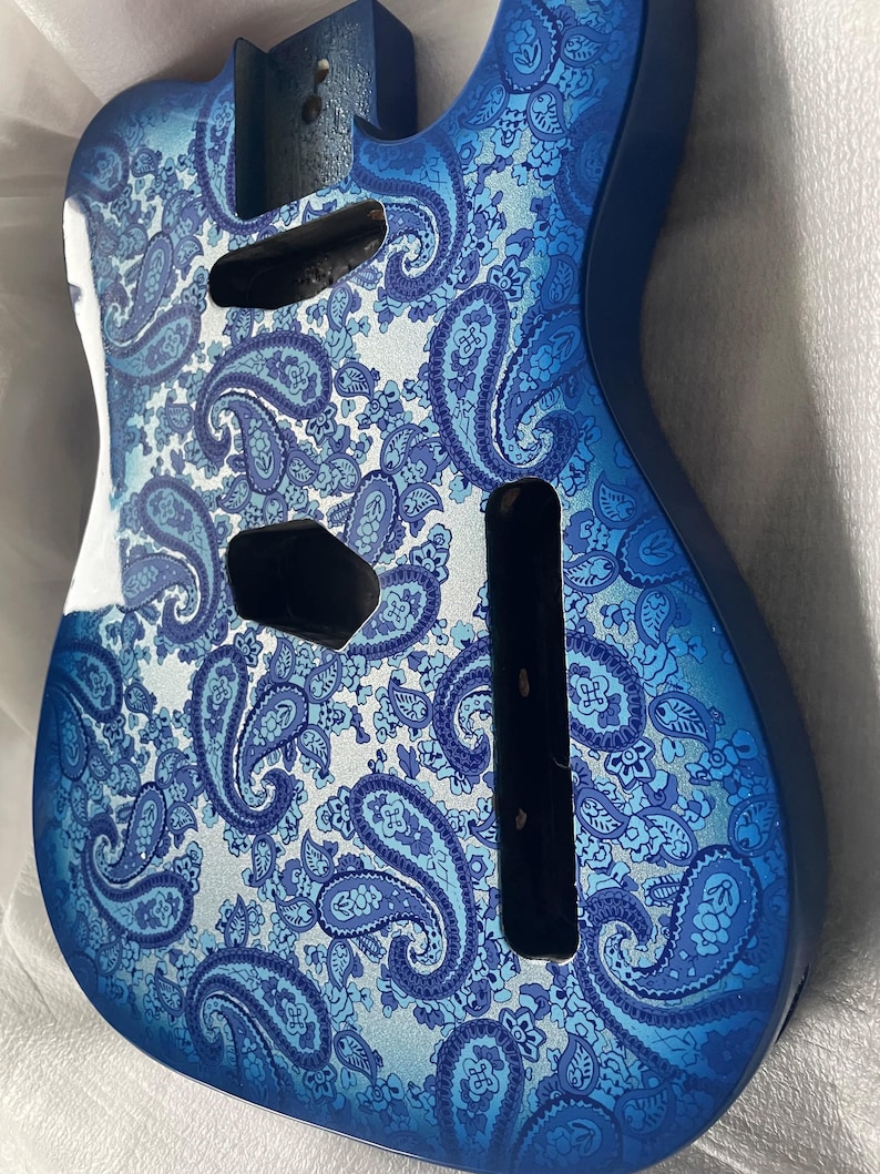 K&ouml;nnte beinhalten: Ein blauer Gitarrenkorpus mit einem Paisley-Muster in Blau- und Wei&szlig;t&ouml;nen. Der Korpus hat Aussparungen f&uuml;r Tonabnehmer und andere Hardware. Die Oberfl&auml;che ist gl&auml;nzend, und das Design ist detailliert.