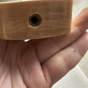 以下が含まれることがあります： 暗い円形のインセットが付いた、明るい色の木製オブジェクトのクローズアップ。 木目は見え、インセットは金属製の継手のようです。 木片に焦点を当てて、手でオブジェクトが保持されています。