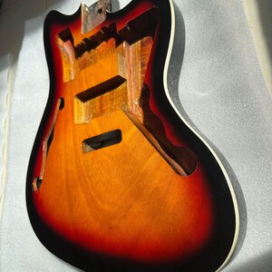 May include: A partially constructed electric guitar body with a sunburst finish. The body features a gradient of colors, transitioning from a dark red to orange and then to a lighter yellow. The edges are black with a white binding.