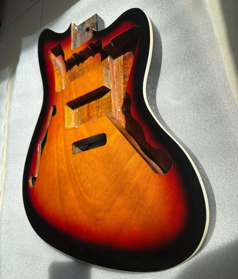 May include: A partially constructed electric guitar body with a sunburst finish. The body features a gradient of colors, transitioning from a dark red to orange and then to a lighter yellow. The edges are black with a white binding.