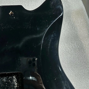May include: A glossy black guitar body with a contoured shape. The body has a smooth, reflective surface with visible scratches and a single hole. The image is a close-up, highlighting the instrument's design and finish.