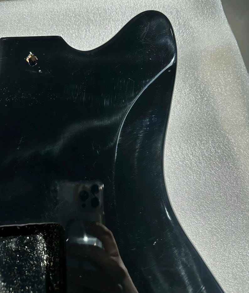 May include: A glossy black guitar body with a contoured shape. The body has a smooth, reflective surface with visible scratches and a single hole. The image is a close-up, highlighting the instrument's design and finish.