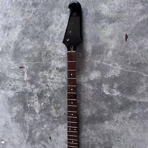 Puede incluir: Un mástil de guitarra negro con un diapasón marrón oscuro. El clavijero es negro brillante con agujeros para las clavijas. El diapasón tiene incrustaciones de puntos blancos. Es una pieza de instrumento musical.