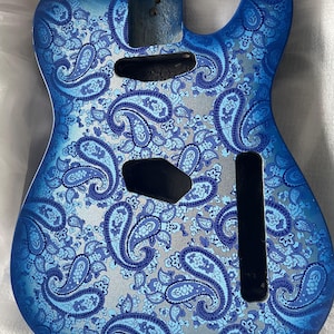 K&ouml;nnte beinhalten: Ein blauer Gitarrenkorpus mit einem Paisley-Muster in Blau- und Silbert&ouml;nen. Er hat Aussparungen f&uuml;r Tonabnehmer und Bedienelemente sowie einen Abschnitt f&uuml;r den Hals. Das Design ist detailliert und aufwendig.