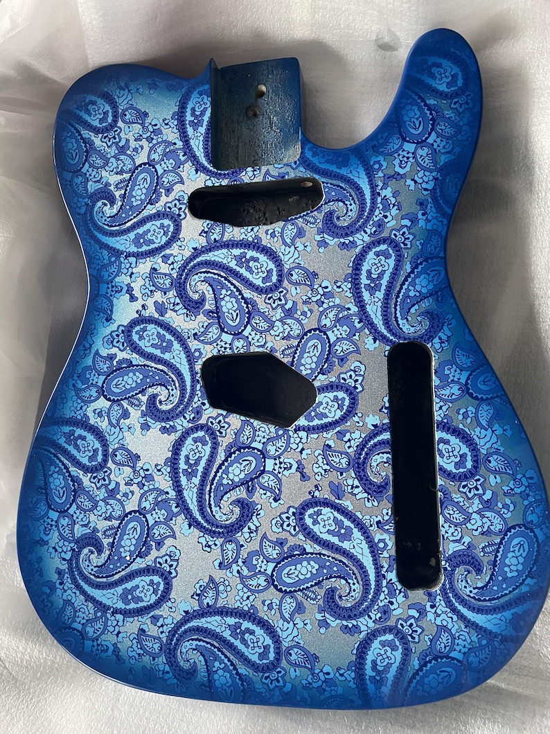 K&ouml;nnte beinhalten: Ein blauer Gitarrenkorpus mit einem Paisley-Muster in Blau- und Silbert&ouml;nen. Er hat Aussparungen f&uuml;r Tonabnehmer und Bedienelemente sowie einen Abschnitt f&uuml;r den Hals. Das Design ist detailliert und aufwendig.