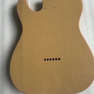 Puede incluir: Un cuerpo de guitarra de color beige claro con forma de Telecaster cl&aacute;sica. El cuerpo tiene un acabado liso y orificios pretaladrados para el m&aacute;stil, el puente y otros componentes. Seis agujeros son visibles cerca del borde inferior para las f&eacute;rulas de las cuerdas.