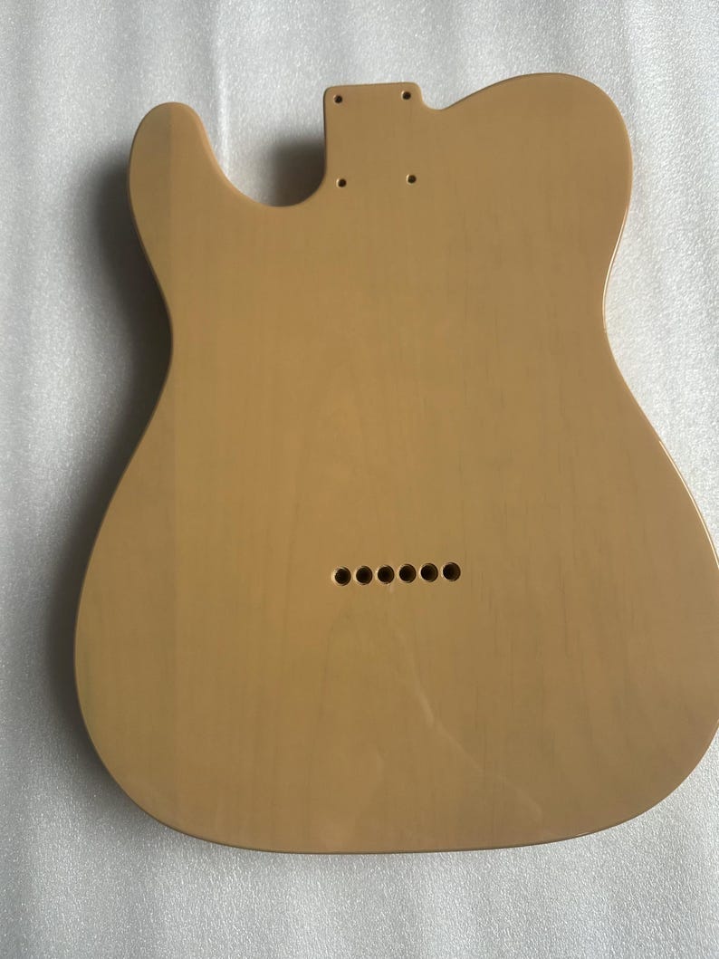 Puede incluir: Un cuerpo de guitarra de color beige claro con forma de Telecaster cl&aacute;sica. El cuerpo tiene un acabado liso y orificios pretaladrados para el m&aacute;stil, el puente y otros componentes. Seis agujeros son visibles cerca del borde inferior para las f&eacute;rulas de las cuerdas.