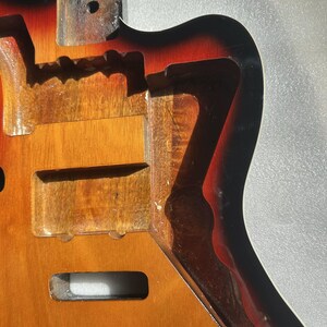 May include: A partially constructed electric guitar body with a sunburst finish. The body is made of wood and features routed cavities for pickups, electronics, and the neck. The finish transitions from yellow to orange to black.