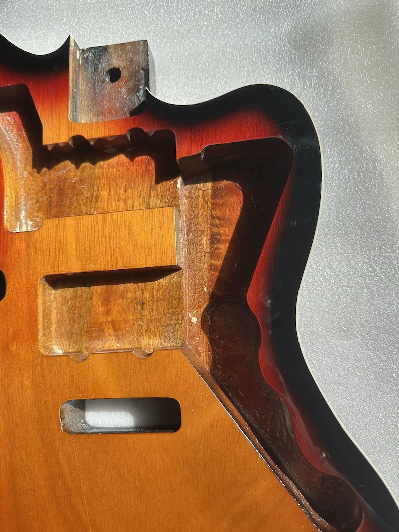 May include: A partially constructed electric guitar body with a sunburst finish. The body is made of wood and features routed cavities for pickups, electronics, and the neck. The finish transitions from yellow to orange to black.