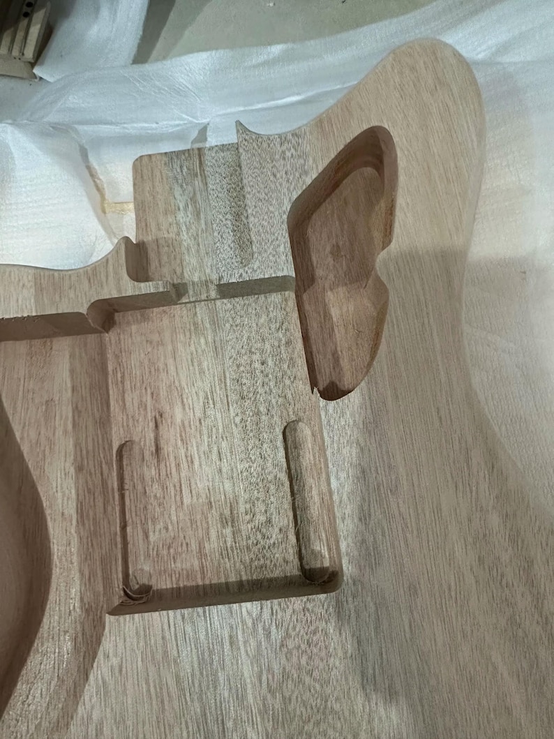 May include: Unfinished wooden guitar body with routed sections for components. The wood grain is visible, showing a light brown color. The body has a curved shape with cutouts for the neck and other hardware.