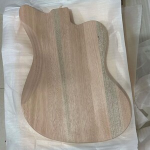 May include: A light-colored, unfinished guitar body made of wood. The body has a contoured shape with a cutaway for access to the higher frets. The wood grain is visible, and the body appears ready for finishing.