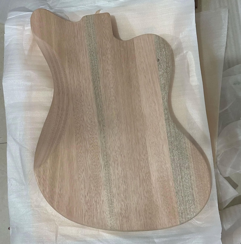 May include: A light-colored, unfinished guitar body made of wood. The body has a contoured shape with a cutaway for access to the higher frets. The wood grain is visible, and the body appears ready for finishing.