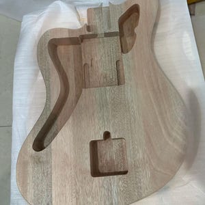 May include: Unfinished wooden guitar body with routed sections for pickups, bridge, and electronics. The natural wood grain is visible, showcasing the instrument's construction. The body is light brown and appears ready for finishing.
