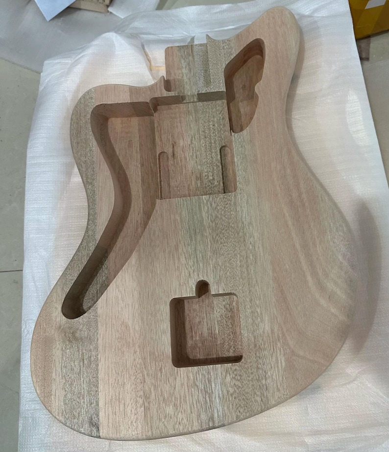 May include: Unfinished wooden guitar body with routed sections for pickups, bridge, and electronics. The natural wood grain is visible, showcasing the instrument's construction. The body is light brown and appears ready for finishing.