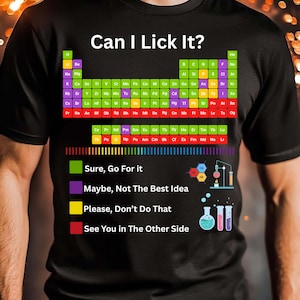 Op de afbeelding: Zwart T-shirt met de tekst "Can I Lick It?" boven een periodiek systeem der elementen. Daaronder kleurgecodeerde waarschuwingen met wetenschappelijke illustraties. Het shirt heeft ook de tekst "DIGITAL PNG FILE".