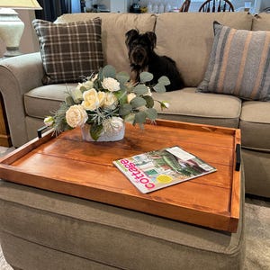 May include: A wooden serving tray with black handles sits on a cushioned ottoman. The tray holds a floral arrangement of white and yellow flowers, and a magazine with the word "cottage" on the cover. A dog sits on the sofa in the background.
