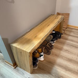 May include: A light-colored wooden shoe bench with a rectangular design. The bench has a natural wood grain and is placed against a neutral-colored wall. Various shoes are stored underneath the bench. The bench is approximately 48 inches long.