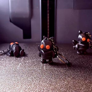 Pode incluir: Três chaveiros de robô pretos com olhos laranja brilhantes são exibidos em uma superfície texturizada. Os chaveiros estão em poses diferentes, com um deitado e os outros em pé. O fundo é de cor escura e neutra.