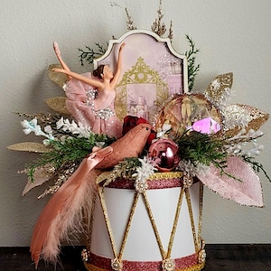 May include: A decorative centerpiece featuring a white drum base with gold glitter trim, topped with a ballerina figurine in a pink dress, a pink bird with a long feather tail, a large faceted crystal, and gold glittered foliage accents.