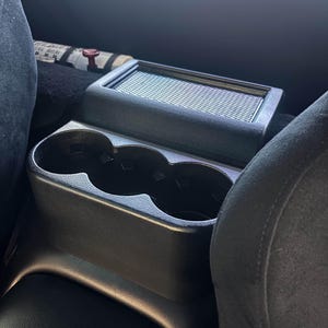 May include: Black car console with two cup holders and a rectangular storage compartment. The console is made of a matte black plastic and is positioned between two black seats.