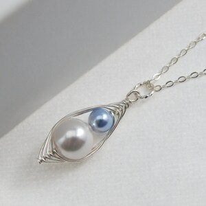 May include: A silver-toned necklace featuring a pendant shaped like a pea pod. The pod holds a large, white pearl and a smaller, light blue pearl. The chain is delicate and silver.