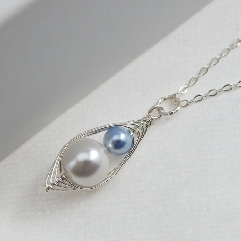May include: A silver-toned necklace featuring a pendant shaped like a pea pod. The pod holds a large, white pearl and a smaller, light blue pearl. The chain is delicate and silver.