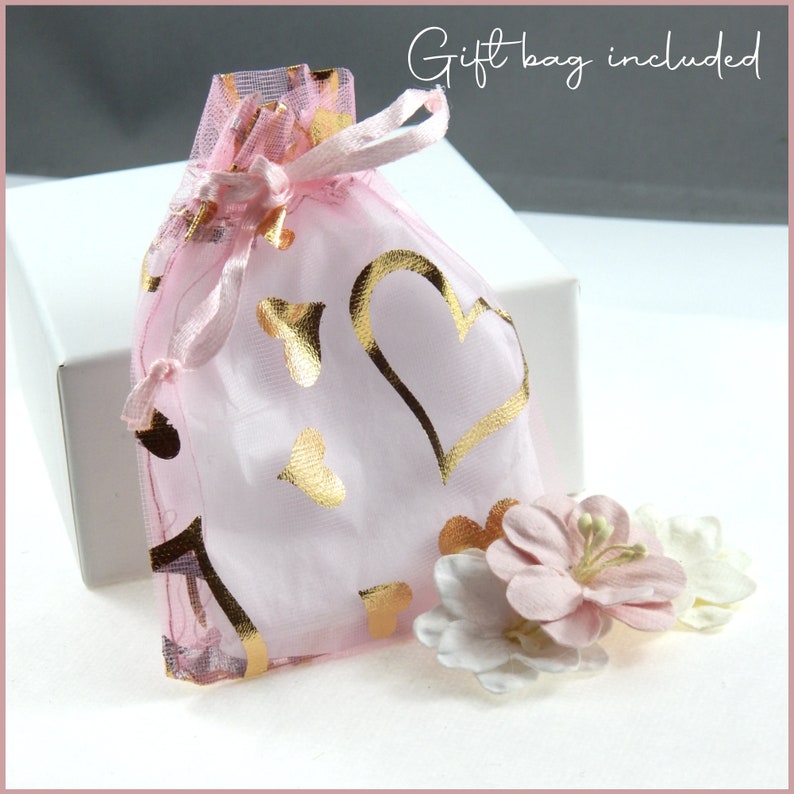 May include: A pink organza gift bag with gold heart and butterfly designs, tied with a matching ribbon. The bag sits in front of a white box, with pink and white fabric flowers nearby. Text reads "Gift bag included."
