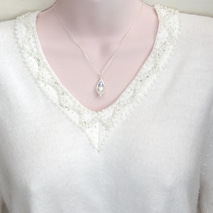 May include: A silver necklace with a teardrop-shaped pendant. The pendant features a white pearl and a blue stone, encased in a delicate silver wire. The necklace is displayed on a white sweater with pearl embellishments.