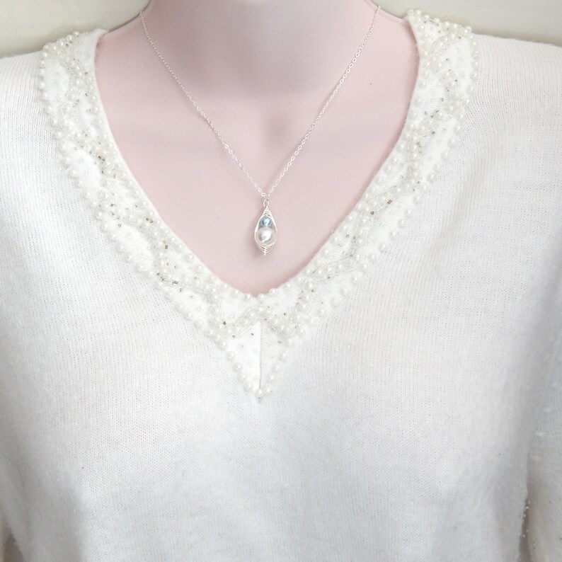 May include: A silver necklace with a teardrop-shaped pendant. The pendant features a white pearl and a blue stone, encased in a delicate silver wire. The necklace is displayed on a white sweater with pearl embellishments.