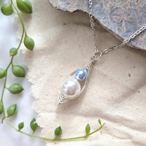 May include: A silver-toned necklace featuring a pendant with a wire-wrapped design. The pendant holds a large white pearl and a smaller blue pearl. The necklace is displayed on a textured, off-white surface, with green plant stems and leaves in the background.