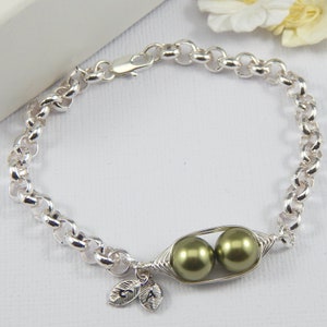 May include: A silver chain bracelet with a charm of two green pearls in a silver pod. The charm has two small silver leaves with the letters "J" and "A" engraved on them.