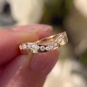 Pode incluir: Um anel de ouro curvo com uma série de pedras preciosas claras redondas e em forma de marquesa. O anel é segurado na mão, com um fundo floral desfocado. A palavra "ECORY" está gravada no anel.