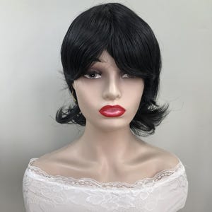 May include: A black wig with short, layered hair and bangs. The wig is styled on a mannequin head with red lipstick and a white lace top. The wig is a dark, solid color and appears to be made of synthetic fibers.