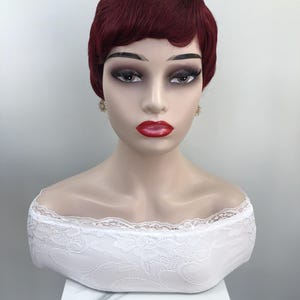May include: A mannequin head with a short, burgundy-colored wig styled with waves. The mannequin has full makeup, including red lipstick and gold starburst earrings. A white lace off-the-shoulder top is visible.