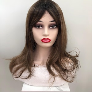 May include: A brown wig with dark roots and long, wavy layers is displayed on a mannequin head. The mannequin has full makeup, including red lipstick. The wig is styled with bangs and a side part. The mannequin is on a white stand.