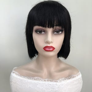 May include: A mannequin head with a black bob wig featuring blunt bangs and a chin-length cut. The mannequin has full makeup, including dark eye shadow and bright red lipstick. The mannequin is wearing a white lace top.