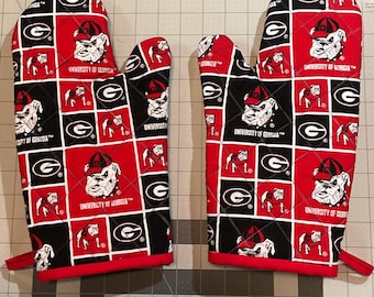 Puede incluir: Un par de manoplas acolchadas para horno con un patrón repetido del logotipo del bulldog de la Universidad de Georgia, la letra "G" y las palabras "University of Georgia" sobre un fondo rojo, negro y blanco. Las manoplas tienen ribetes rojos.