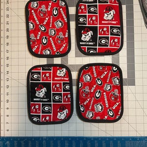 May include: Four rectangular pot holders with black edging. Two feature a red background with white and black UGA Bulldogs logos and text. The other two have a red, black, and white checkerboard pattern with the University of Georgia Bulldogs logo.