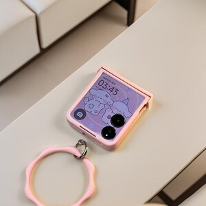 May include: A pink foldable smartphone with a matching pink ring holder. The phone screen displays a cartoon design and the time 03:43. The phone is on a light-colored surface.