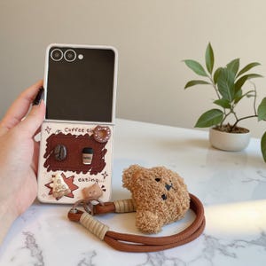 May include: A white and brown phone case with a coffee theme, featuring a bear-shaped strap. The case has a brown interior with coffee-related charms and the text "Coffee time eating...". A fluffy brown bear head is attached to the strap.
