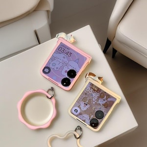 May include: Pink and beige phone cases with cartoon designs and a small cat figurine on top. One case displays the time 03:43, and the other 03:22. A pink, scalloped-edge accessory and a white strap are also visible.