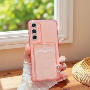 May include: A clear pink phone case with a cardholder. The case has a transparent design with a pink border and a clear card slot on the back. The phone case has three camera lenses. The word "SAMSUNG" is visible on the cardholder.
