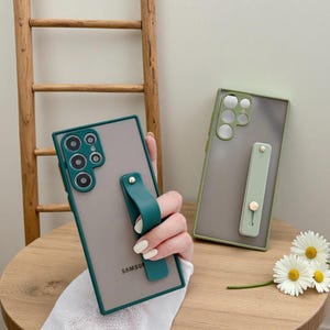 May include: Two phone cases with a clear back and coloured frame. One is teal with a matching grip, the other is olive green with a light green grip. The cases have a Samsung logo.