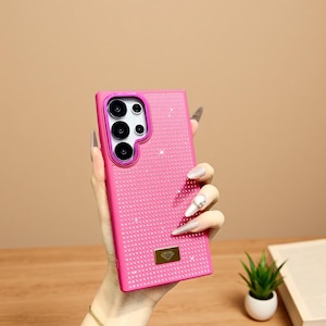 May include: A bright pink phone case with a textured surface and a decorative diamond pattern. The case has a gold-colored emblem and a camera cutout with a pink border. The phone case is designed to protect the phone.