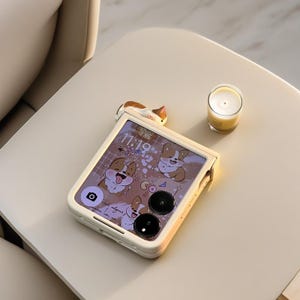 May include: A cream-colored phone case with a cartoon corgi design. The phone case is open, displaying the phone screen with the time 11:19. A small candle in a glass and a corgi-shaped charm are also visible.