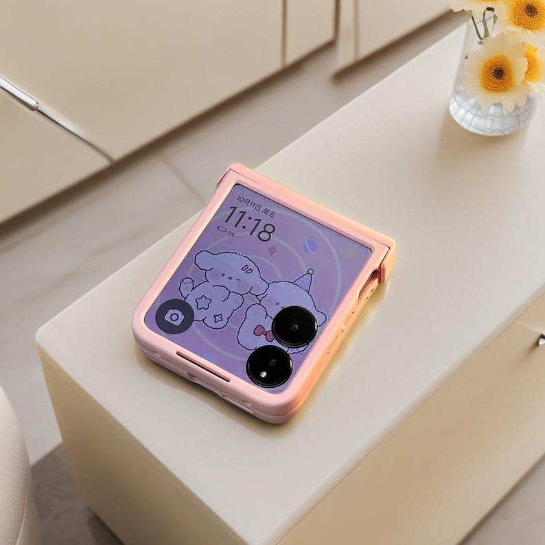May include: A pink smartphone with a cartoon graphic on the screen, displaying the time and date. The phone is in a pink case and rests on a white surface. A vase of flowers is in the background.