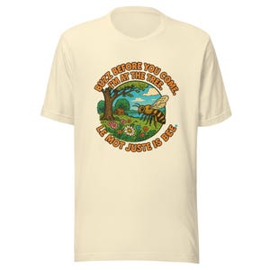 May include: Cream-colored t-shirt featuring a circular graphic with a bee, tree, and flowers. The text reads "Buzz before you come, I'm at the tree. Le mot juste is bee."