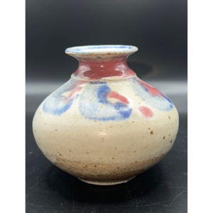 Puede incluir: Un pequeño jarrón de cerámica hecho a mano con un cuerpo redondeado y un cuello estrecho. El jarrón presenta una base de color crema con diseños abstractos azules y rojos. El borde es de color rojo intenso y el jarrón tiene una estética rústica y terrosa.