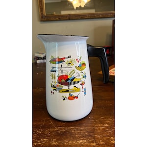 May include: White enamelware pitcher with a black handle and rim. The pitcher features a colorful, vintage-style illustration of kitchen-themed objects. The pitcher is a classic kitchenware item.